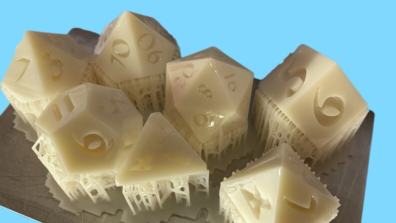 Pode incluir: Um conjunto de dados de v&aacute;rias faces em branco sujo para jogos de tabuleiro. Os dados t&ecirc;m v&aacute;rias formas geom&eacute;tricas, com n&uacute;meros gravados nas suas superf&iacute;cies. Parecem rec&eacute;m-sa&iacute;dos de uma impressora 3D, com estruturas de suporte ainda presas.