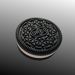 Pode incluir: Close-up de um biscoito Oreo. O biscoito preto tem um padr&atilde;o detalhado e a palavra "OREO" no centro. O recheio de creme branco &eacute; vis&iacute;vel entre as duas metades do biscoito. O biscoito est&aacute; em uma superf&iacute;cie reflexiva.