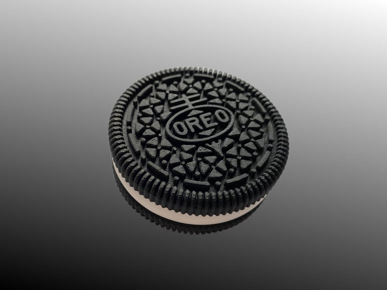 Pode incluir: Close-up de um biscoito Oreo. O biscoito preto tem um padr&atilde;o detalhado e a palavra "OREO" no centro. O recheio de creme branco &eacute; vis&iacute;vel entre as duas metades do biscoito. O biscoito est&aacute; em uma superf&iacute;cie reflexiva.