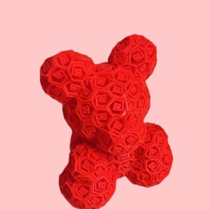 Pode incluir: Um objeto vibrante em forma de urso vermelho com um padr&atilde;o geom&eacute;trico. O urso tem uma superf&iacute;cie texturizada com um design hexagonal repetitivo. O fundo &eacute; rosa suave, criando um contraste marcante com a cor do urso.