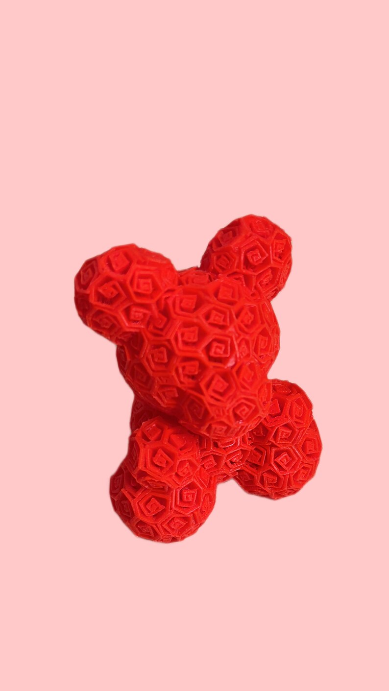 Pode incluir: Um objeto vibrante em forma de urso vermelho com um padr&atilde;o geom&eacute;trico. O urso tem uma superf&iacute;cie texturizada com um design hexagonal repetitivo. O fundo &eacute; rosa suave, criando um contraste marcante com a cor do urso.