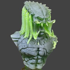 Pode incluir: Um busto de Predador verde transl&uacute;cido e preto. A escultura apresenta uma armadura detalhada, um rosto amea&ccedil;ador com dentes afiados e rastas verdes distintas. O busto est&aacute; sobre um fundo cinza escuro.