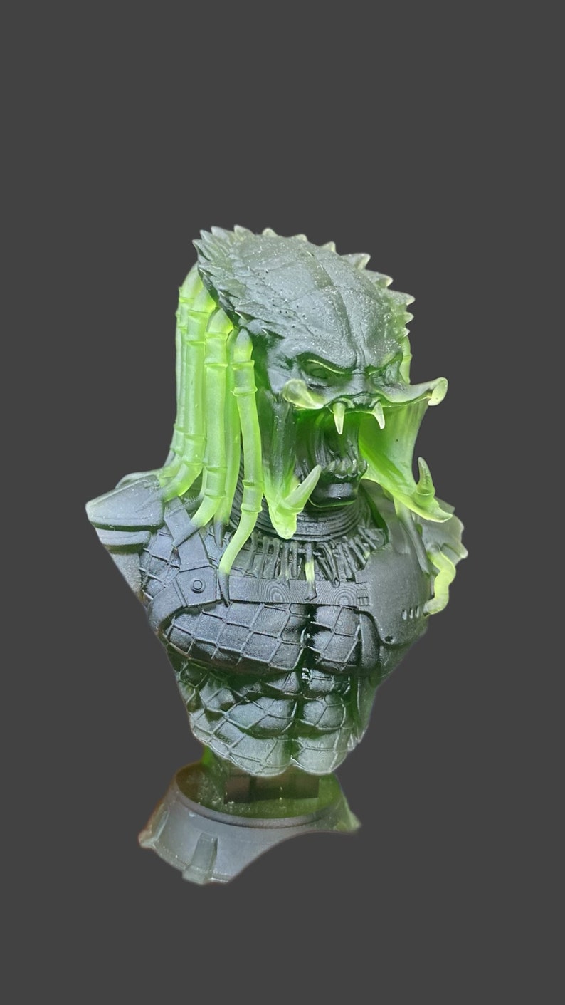 Pode incluir: Um busto de Predador verde transl&uacute;cido e preto. A escultura apresenta uma armadura detalhada, um rosto amea&ccedil;ador com dentes afiados e rastas verdes distintas. O busto est&aacute; sobre um fundo cinza escuro.