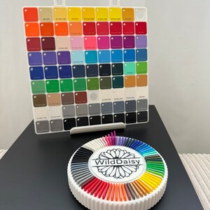 Pode incluir: Um organizador de amostras de cores branco com uma grade de pequenos quadrados coloridos. Abaixo, um suporte circular branco exibe um espectro de hastes coloridas, com "WildDaisy" impresso no centro.
