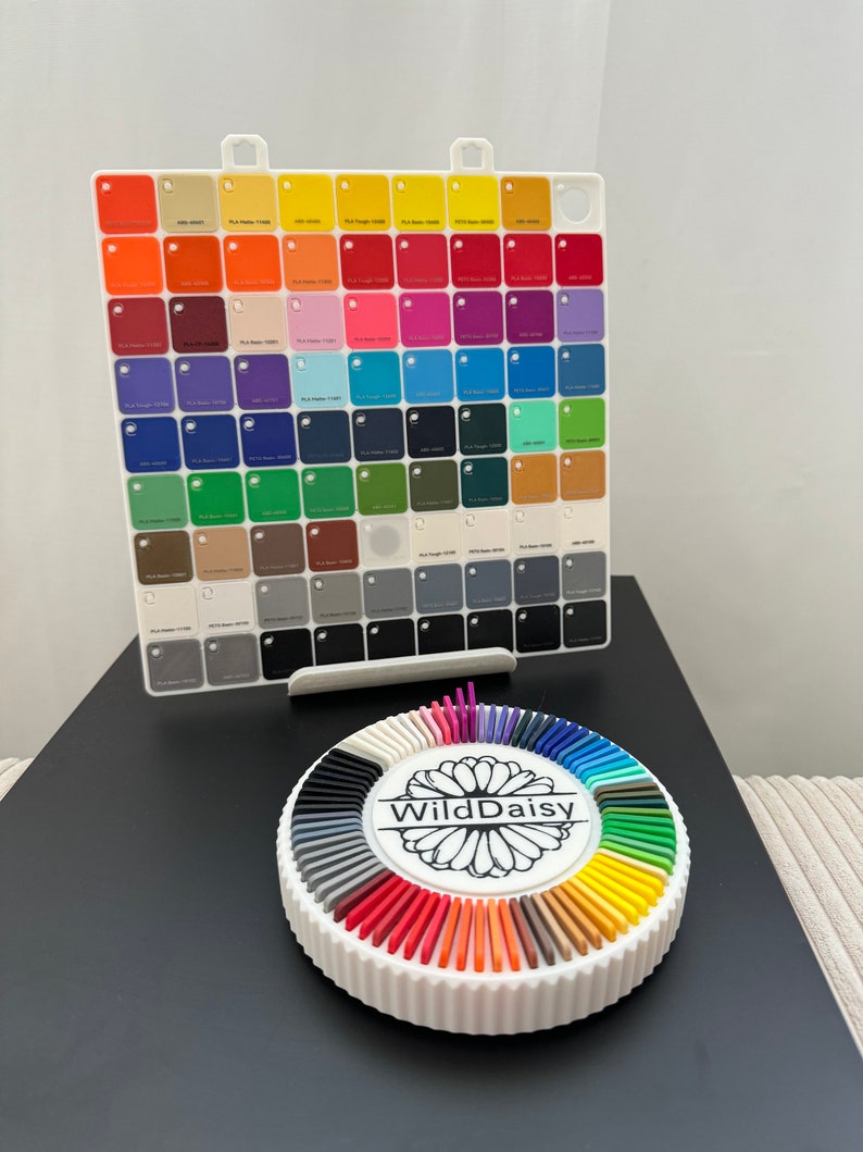 Pode incluir: Um organizador de amostras de cores branco com uma grade de pequenos quadrados coloridos. Abaixo, um suporte circular branco exibe um espectro de hastes coloridas, com "WildDaisy" impresso no centro.