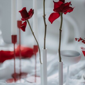 Pode incluir: Uma natureza morta rom&acirc;ntica com tr&ecirc;s rosas vermelhas escuras em vasos cil&iacute;ndricos brancos. Velas brancas altas em suportes vermelhos e um tecido de cetim vermelho criam um ambiente rom&acirc;ntico e desfocado. O arranjo &eacute; elegante.