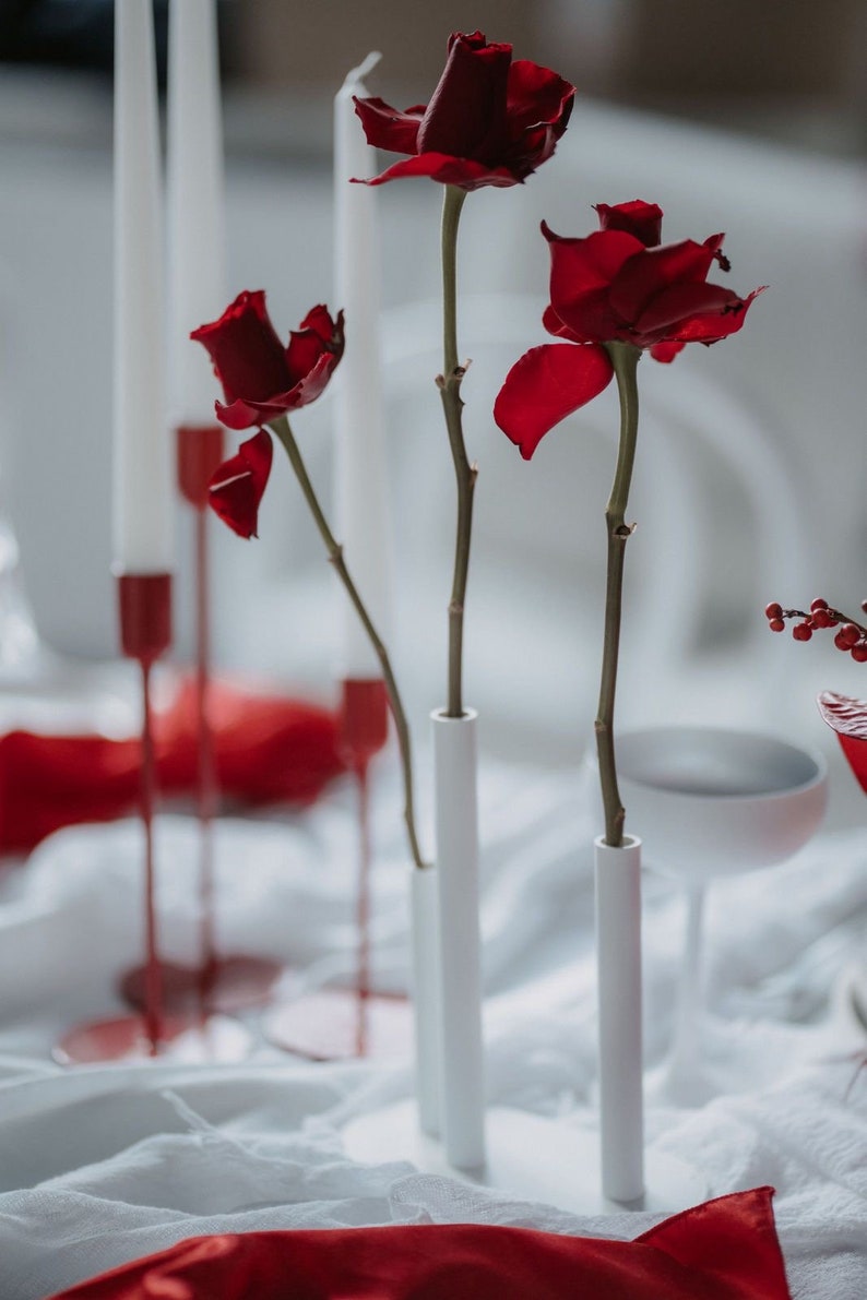 Pode incluir: Uma natureza morta rom&acirc;ntica com tr&ecirc;s rosas vermelhas escuras em vasos cil&iacute;ndricos brancos. Velas brancas altas em suportes vermelhos e um tecido de cetim vermelho criam um ambiente rom&acirc;ntico e desfocado. O arranjo &eacute; elegante.