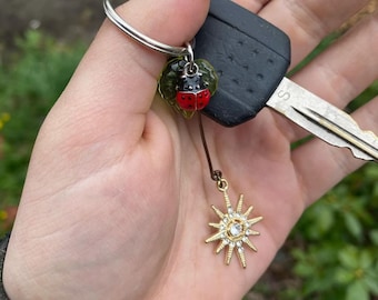 May include: A gold star charm with rhinestones hangs from a key chain with a ladybug charm. The ladybug charm is red with black spots and is attached to a green leaf.