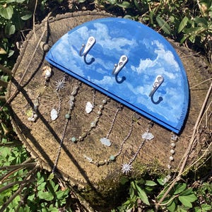 May include: A blue wooden half-circle wall hook with a painted sky and clouds design. Three silver hooks are attached to the top. A chain of charms hangs from the bottom, including clouds, stars, and beads.