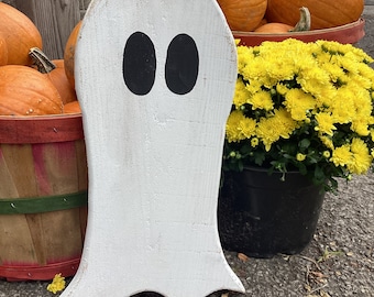 Puede incluir: Un recorte de fantasma de madera blanca con ojos pintados de negro. El fantasma está de pie sobre una superficie pavimentada frente a una cesta de calabazas naranjas y crisantemos amarillos.