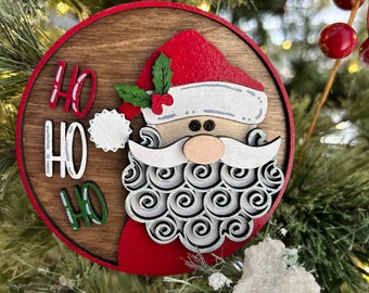 May include: A round wooden Christmas ornament with a red and white Santa Claus face and the words "Ho Ho Ho" in green and white lettering.