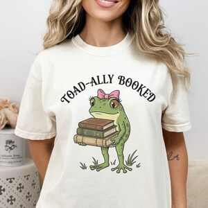 May include: Ivory-colored t-shirt featuring a cartoon frog holding books, with the text "TOAD-ALLY BOOKED" above. The frog is green with a pink bow. The shirt is a relaxed fit.