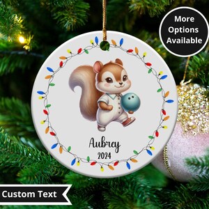 May include: A round Christmas ornament featuring a cartoon squirrel holding a bowling ball. The squirrel is wearing a white outfit. The ornament is decorated with a string of colorful lights and the name "Aubrey" and the year "2024".