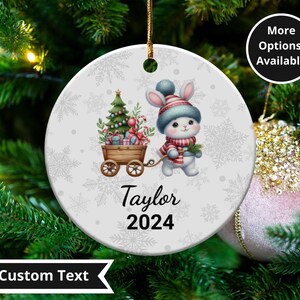 May include: A round Christmas ornament with a winter theme. It features a cartoon rabbit wearing a hat and scarf, pulling a wooden cart with a Christmas tree and gifts. The name "Taylor" and the year "2024" are printed on the ornament.