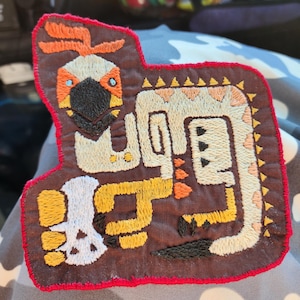 Pode incluir: Patch bordado com um desenho de criatura estilizada nas cores laranja, amarelo e creme sobre fundo castanho. Possui uma borda vermelha e um detalhe de caveira. O design é detalhado e intrincado, para roupas ou acessórios.