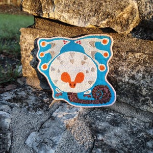 May include: Embroidered patch featuring a stylized creature design with a white body, orange beak, and blue accents. The patch has a light blue border and a brown spiral detail. The patch is placed on a stone surface.
