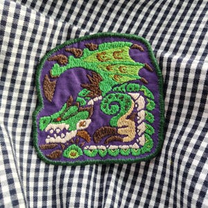 Könnte beinhalten: Ein gestickter Aufnäher mit einem stilisierten grünen Drachen mit braunen Akzenten auf violettem Grund. Der Drache hat einen sichtbaren Flügel, einen langen Schwanz und spitze Zähne. Das Design ist mit einem dunkelgrünen Rand umstickt.