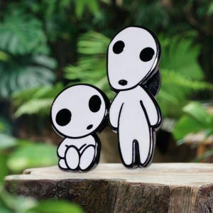 May include: Two enamel pins featuring white figures with black eyes and outlines. One figure is seated, the other standing. The pins are set against a blurred green background, suggesting a nature theme.