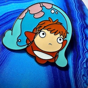 May include: A red and blue enamel pin featuring a cartoon character with red hair and large eyes. The character is wearing a red and white outfit and is surrounded by a blue and pink cloud-like shape.