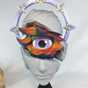 May include: A 3D printed sculpture of a human head with a single eye. The eye is surrounded by a colorful, abstract design. The head is broken into pieces, and the eye is visible through the cracks. The head is on a black stand.