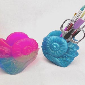May include: Two seashell-shaped desk organizers. One is a gradient of pink and blue, the other is blue and holds scissors and paintbrushes. The organizers have a spiral design and are suitable for storing art supplies.