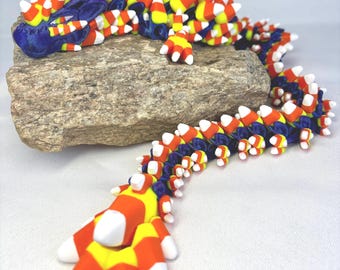 May include: A colorful, articulated dragon figurine with a blue head and body, adorned with orange, yellow, and white candy corn-shaped segments. The dragon rests on a textured, gray stone, with its tail extending downwards.