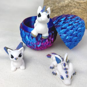 May include: A fantasy-themed set featuring a blue, purple, and pink dragon egg-shaped container with a unicorn figurine. Additional figurines include a white cat-like creature and a white dragon with blue accents. The egg's lid is open.