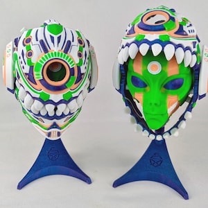 May include: Two futuristic alien masks, one showing the interior. The masks are white with green, orange, and blue accents. They have a detailed design with a large eye-like feature and are mounted on blue stands. The interior mask has a green face.