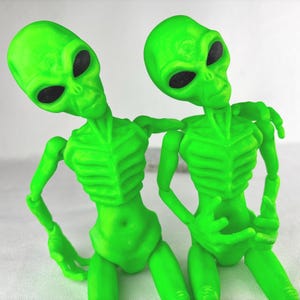 May include: Two green alien figurines with large black eyes. The aliens have slender bodies and articulated limbs. They are posed together, one with an arm around the other. The figures are made of a solid, smooth material.