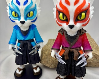 May include: Two figurines of cat-like characters with white faces and detailed facial markings. One wears blue and black clothing, the other red and black. Both hold silver claw-like weapons and wear traditional footwear. The figurines are on small bases.