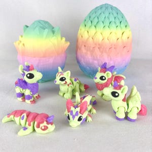 May include: A collection of colorful, miniature dragon figurines and two pastel-colored, geometric egg-shaped containers. The dragons are primarily lime green with purple, pink, and red accents. The eggs are rainbow-colored.