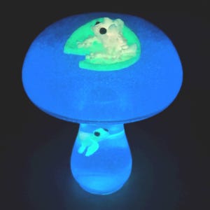 May include: A glowing blue mushroom-shaped night light with a frog theme. The mushroom cap is a bright blue, with a frog on a lily pad. The stem has a frog inside. The frogs and lily pad glow green.