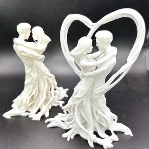 May include: Two white figurines depicting a couple embracing, with one figure framed by a large, sculpted heart shape. Both statues feature flowing, root-like textures at the base, creating a unified appearance of two people dancing together in white.