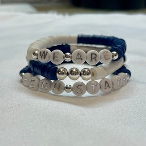 May include: A stack of three beaded bracelets featuring navy blue and white clay discs, interspersed with metallic silver beads. The bracelets display the white letter beads spelling out the phrase WE ARE PENN STATE in silver lettering.
