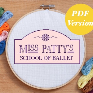 Pode incluir: Um aro de bordado branco com uma placa rosa que diz "Miss Patty's School of Ballet". A placa é cercada por uma borda roxa e tem um pequeno desenho floral na parte superior central. O aro é cercado por fio de bordado colorido.