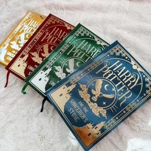 May include: Four hardcover books titled Harry Potter and the Sorcerer's Stone, each featuring gold foil illustrations of a castle, an owl, and a crescent moon. The covers are yellow, red, green, and blue, with matching ribbon bookmarks attached.