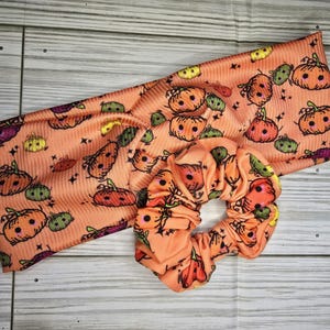 May include: An orange headband and scrunchie set with a repeating pattern of cartoon pumpkins in various colors. The pumpkins have black outlines and are set against a ribbed fabric.