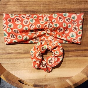 Op de afbeelding: Een rood en wit kerstthema hoofdband met een bijpassende scrunchie. Het hoofdband en de scrunchie hebben een herhalend patroon van kleine rode cirkels met witte omtrekken, elk met een ander kerstpictogram, zoals een sneeuwpop, een rendier, een kerstboom en een kerstmanmuts.