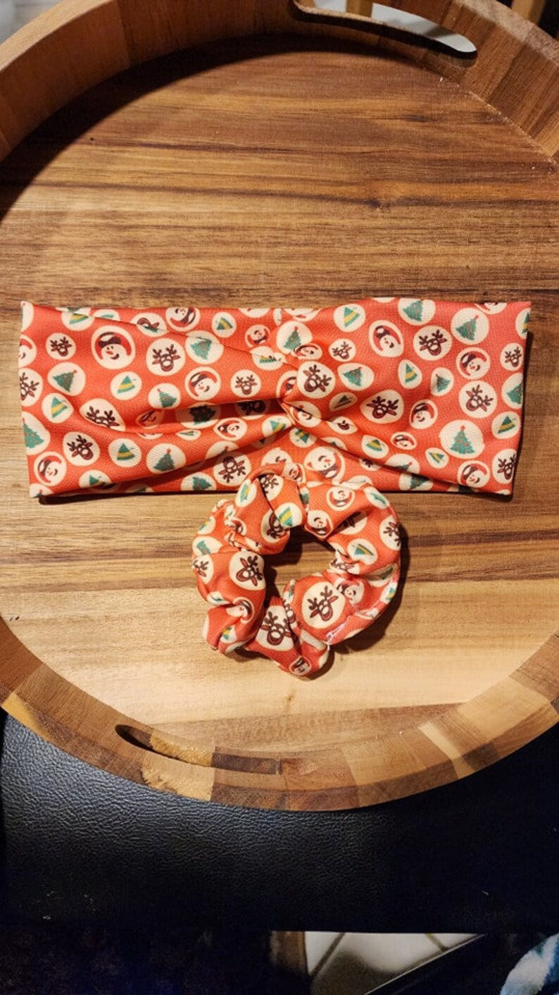 Op de afbeelding: Een rood en wit kerstthema hoofdband met een bijpassende scrunchie. Het hoofdband en de scrunchie hebben een herhalend patroon van kleine rode cirkels met witte omtrekken, elk met een ander kerstpictogram, zoals een sneeuwpop, een rendier, een kerstboom en een kerstmanmuts.