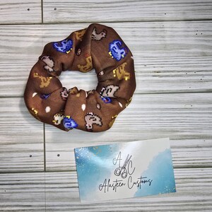 Pu&ograve; includere: Un elastico per capelli marrone con un motivo ripetuto di piccoli animali pixelati in marrone, giallo e blu. L'elastico &egrave; su una superficie di legno. Il testo "A.C. Alasteen Customs" &egrave; su un cartoncino blu e bianco.