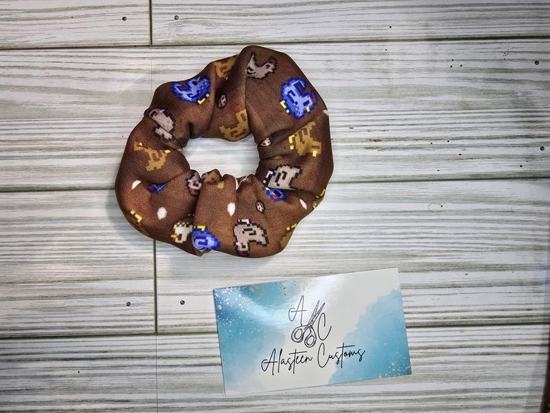 Pu&ograve; includere: Un elastico per capelli marrone con un motivo ripetuto di piccoli animali pixelati in marrone, giallo e blu. L'elastico &egrave; su una superficie di legno. Il testo "A.C. Alasteen Customs" &egrave; su un cartoncino blu e bianco.