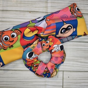 Op de afbeelding: Een stoffen hoofdband en een bijpassende scrunchie met een kleurrijk, herhalend patroon van animatiefiguren, waaronder oranje anemoonvissen, een gele bal met een rode ster en een baby met een zwart masker op roze, blauwe en gele blokken.