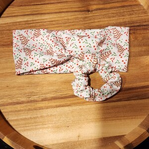 Op de afbeelding: Een wit hoofdband met een kerstthema print van rode en groene pepermuntstokken en sterren op een witte achtergrond. Het hoofdband is in een knoop geknoopt en heeft een bijpassende scrunchie.