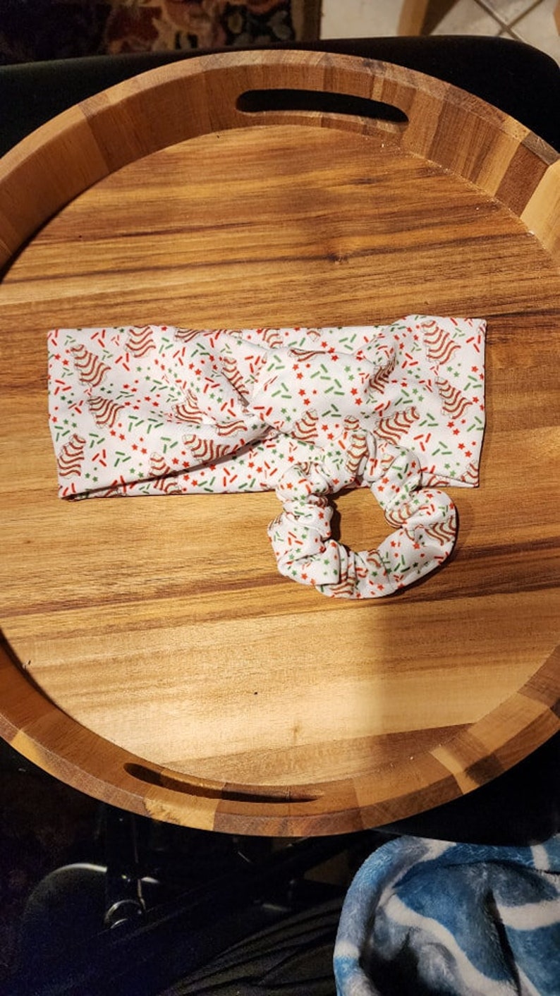 Op de afbeelding: Een wit hoofdband met een kerstthema print van rode en groene pepermuntstokken en sterren op een witte achtergrond. Het hoofdband is in een knoop geknoopt en heeft een bijpassende scrunchie.