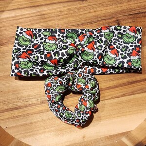Op de afbeelding: Een set van twee Grinch-thema haaraccessoires. Een zwart-wit luipaardprint hoofdband met een groen Grinch-gezicht en een rood kerstman-hoedontwerp. Een bijpassende scrunchie met hetzelfde ontwerp.