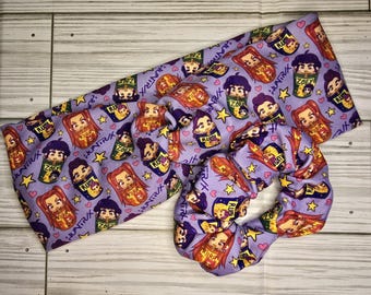 Può includere: Un set di fascia e scrunchie in tessuto lavanda. Il tessuto presenta un motivo ripetuto di personaggi dei cartoni animati, stelle e cuori. È visibile anche il testo "HUNTRAX" e "ZOEY". Lo scrunchie è un grande cerchio arricciato.