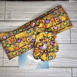 May include: A yellow fabric headband with a repeating pattern of small pixelated characters in various colors. The headband is paired with a matching yellow scrunchie with the same pattern. The text "Alasten Customs" is printed on a white card in the foreground.