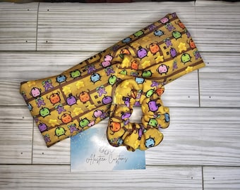 Può includere: Un cerchietto per capelli in tessuto giallo con un motivo ripetuto di piccoli personaggi pixelati in vari colori. Il cerchietto è abbinato a un elastico per capelli giallo abbinato con lo stesso motivo. Il testo "Alasten Customs" è stampato su una carta bianca in primo piano.