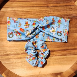 Op de afbeelding: Een blauwe hoofdband en scrunchie set met een kerstthema print met een sneeuwpop, een Grinch-achtig personage en cadeaus. De print is op een lichtblauwe achtergrond.
