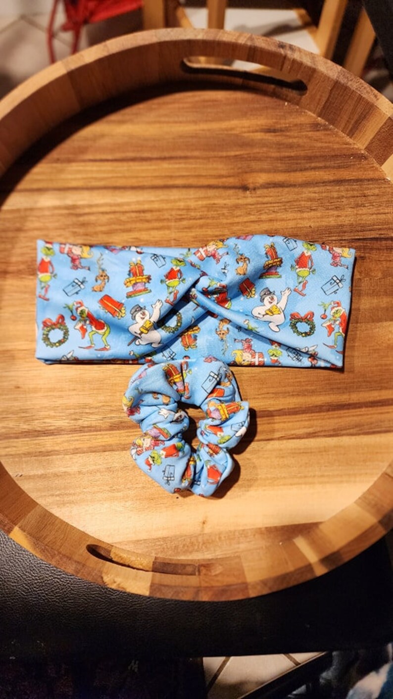 Op de afbeelding: Een blauwe hoofdband en scrunchie set met een kerstthema print met een sneeuwpop, een Grinch-achtig personage en cadeaus. De print is op een lichtblauwe achtergrond.